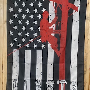 Op de afbeelding: Een Amerikaanse vlag met een rode streep aan de rechterkant. In het midden van de vlag staat een silhouet van een persoon die een elektriciteitspaal beklimt. Onderaan de vlag staan vijf gereedschappen: een tang, een schroevendraaier, een moersleutel, een tang en een hamer.