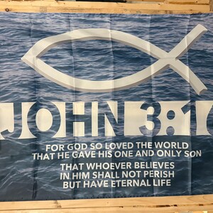 May include: A flag with a blue water background and a white fish symbol. The text "JOHN 3:16" is prominently displayed, along with a quote about faith and eternal life. The flag is rectangular and has grommets.