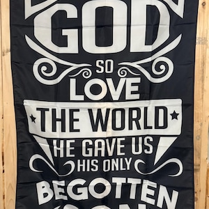 May include: Black flag with white text. The text reads "FOR GOD SO LOVE THE WORLD HE GAVE US HIS ONLY BEGOTTEN SON JOHN 3:16". The flag has a decorative design with swirls and stars.