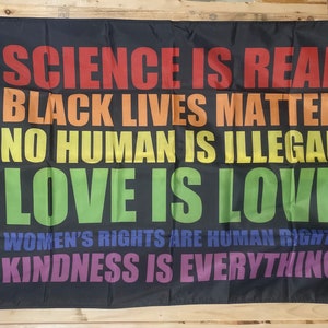 May include: A black flag with rainbow colored text that reads "SCIENCE IS REAL", "BLACK LIVES MATTER", "NO HUMAN IS ILLEGAL", "LOVE IS LOVE", "WOMEN'S RIGHTS ARE HUMAN RIGHTS", and "KINDNESS IS EVERYTHING".