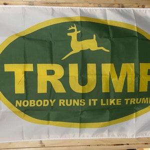 May include: A white flag with a green oval and yellow outline. Inside the oval is a yellow deer silhouette and the word "TRUMP" in yellow letters. Below the word is the text "NOBODY RUNS IT LIKE TRUMP".