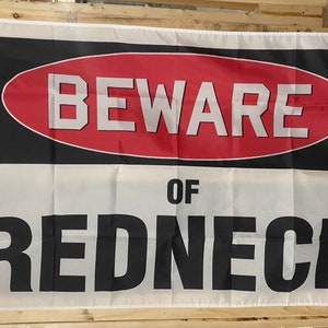 Könnte beinhalten: Eine weiße Flagge mit schwarzem Text, der "BEWARE OF REDNECK" in einem roten und schwarzen ovalen Warnschild lautet.