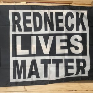 Könnte beinhalten: Schwarze Flagge mit weißem Text, der "REDNECK LIVES MATTER" in einer fetten, blockartigen Schriftart zeigt. Der Text ist in drei Zeilen angeordnet, wobei jedes Wort in einem weißen Rechteck zentriert ist. Die Flagge hat Ösen zum Aufhängen.