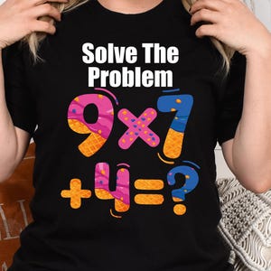Op de afbeelding: Zwart T-shirt met de tekst "Solve The Problem" boven een kleurrijke wiskundige vergelijking: 9 x 7 + 4 = ?. De getallen en symbolen zijn in heldere kleuren, waaronder roze, oranje en blauw. Het shirt is een casual stijl.