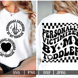 Peut inclure: Sweat-shirt blanc avec un texte noir qui dit "PERSONALLY VICTIMIZED BY MY TODDLER" dans une police rétro. Le texte est répété deux fois, une fois avec un signe de paix et un graphique de main de squelette et une fois avec un graphique de cœur qui coule.