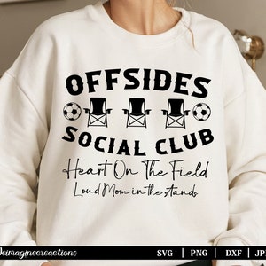 Op de afbeelding: Wit sweatshirt met zwarte tekst die "OFFSIDES SOCIAL CLUB Heart On The Field Loud Mom in the stands" luidt. De tekst wordt omringd door drie zwarte tuinstoelen en twee zwart-witte voetballen.