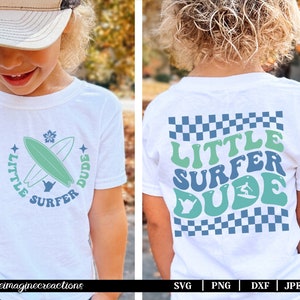 Puede incluir: Camiseta blanca con un diseño gráfico azul y verde que dice "Little Surfer Dude" con una tabla de surf y una ola. El diseño es de estilo retro.