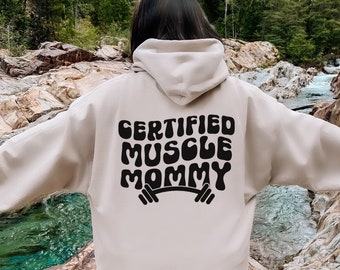 Może przedstawiać: Beżowa bluza z kapturem z czarnym nadrukiem "CERTIFIED MUSCLE MOMMY" z sztangą pod spodem.