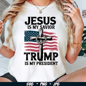 May include: White t-shirt with a red, white and blue American flag featuring a cross and the text "Jesus is my Saviour, Trump is my President".