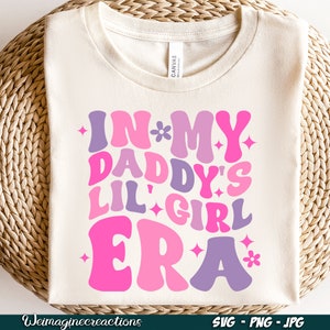 Peut inclure: Un t-shirt blanc avec un motif rose, violet et blanc qui dit "In My Daddy's Lil' Girl Era" avec des &eacute;toiles et des fleurs.