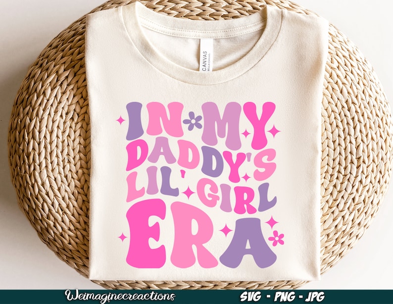 Peut inclure: Un t-shirt blanc avec un motif rose, violet et blanc qui dit "In My Daddy's Lil' Girl Era" avec des &eacute;toiles et des fleurs.
