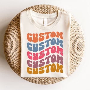 May include: A white t-shirt with the word "CUSTOM" repeated five times in a retro, colorful font. Each "CUSTOM" is a different color: orange, blue, pink, purple, and yellow.