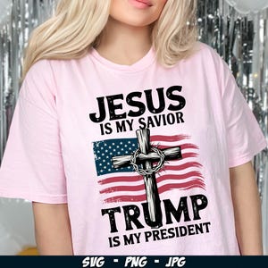 May include: Pink t-shirt with a graphic design featuring a cross with a crown of thorns, an American flag, and the text "Jesus is my Saviour, Trump is my President".