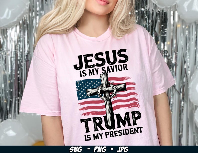 May include: Pink t-shirt with a graphic design featuring a cross with a crown of thorns, an American flag, and the text "Jesus is my Saviour, Trump is my President".