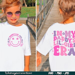 Peut inclure: T-shirt blanc avec un motif rose et violet qui dit "In My Daddy's Lil' Girl Era" avec un smiley sur le devant et un motif similaire au dos.