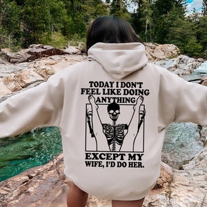 May include: A beige hoodie with a black graphic print of a skeleton doing a yoga pose. The text on the hoodie reads "Today I don't feel like doing anything except my wife, I'd do her."