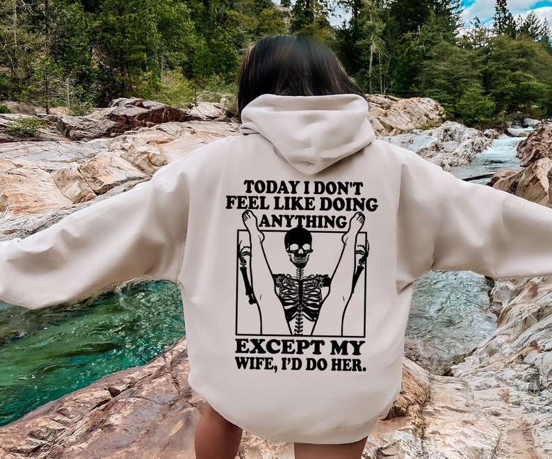 May include: A beige hoodie with a black graphic print of a skeleton doing a yoga pose. The text on the hoodie reads "Today I don't feel like doing anything except my wife, I'd do her."