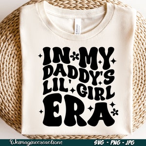 Peut inclure: T-shirt blanc avec du texte noir qui dit "In My Daddy's Lil' Girl Era" avec des &eacute;toiles et des fleurs autour du texte.