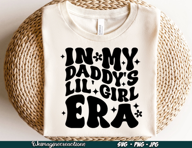 Peut inclure: T-shirt blanc avec du texte noir qui dit "In My Daddy's Lil' Girl Era" avec des &eacute;toiles et des fleurs autour du texte.