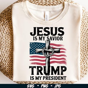 May include: White t-shirt featuring a red, white and blue American flag design. The flag incorporates a wooden cross with thorns. The text "Jesus is my Savior Trump is my President" is printed on the shirt.
