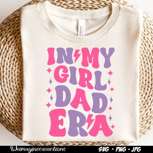 May include: A white t-shirt with the text "In My Girl Dad Era" in pink and purple letters with a lightning bolt and stars.