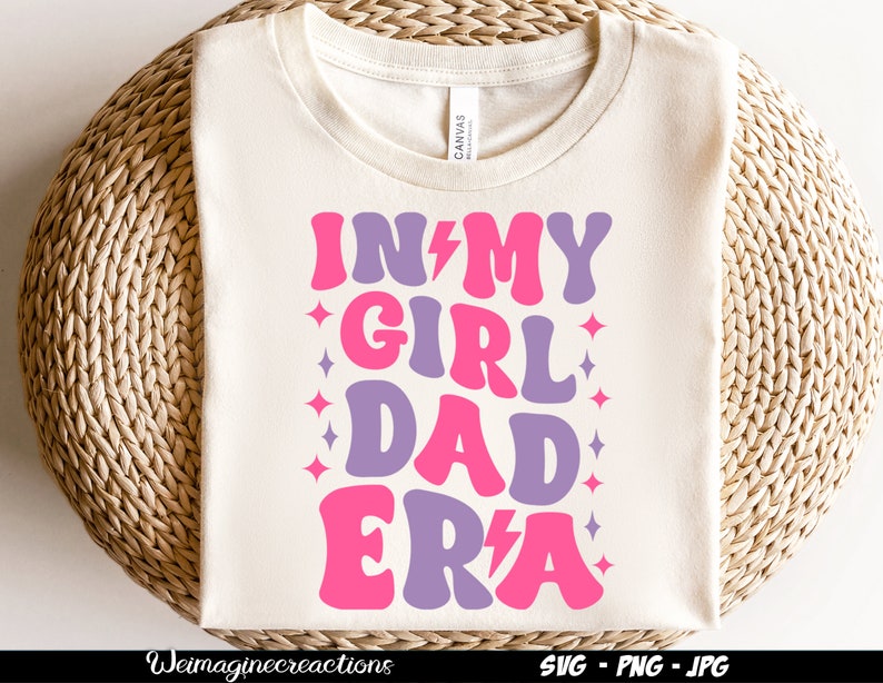 May include: A white t-shirt with the text "In My Girl Dad Era" in pink and purple letters with a lightning bolt and stars.