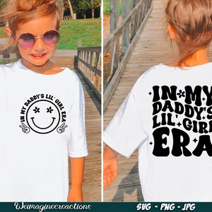 Peut inclure: T-shirt blanc avec un motif noir qui dit "In my Daddy's Lil' Girl Era" avec un smiley avec des fleurs pour les yeux sur le devant et le m&ecirc;me texte dans une police r&eacute;tro au dos.
