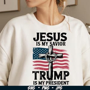 May include: White sweatshirt with a graphic design featuring a cross with a crown of thorns, an American flag, and the text "Jesus is my Saviour Trump is my President".