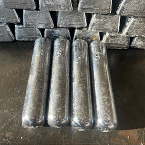 May include: Four silver-colored fishing sinkers, each marked with the number "33", are arranged in a row on a dark gray surface. The sinkers are cylindrical with rounded ends.