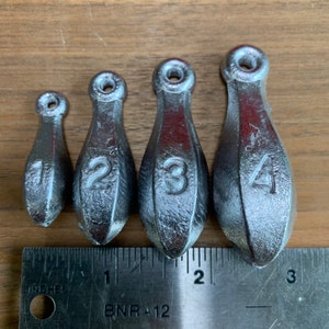 May include: Four silver fishing sinkers, each with a different number from 1 to 4, are arranged in a row on a wooden surface. The sinkers are teardrop-shaped and have a small loop at the top.