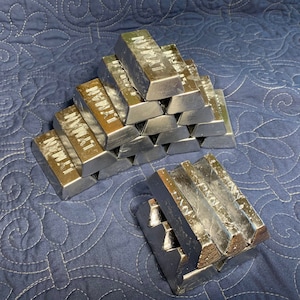 May include: A stack of silver-colored metal bars with the word "LYMAN" printed on them. The bars are arranged in a pyramid shape on a blue quilted surface.
