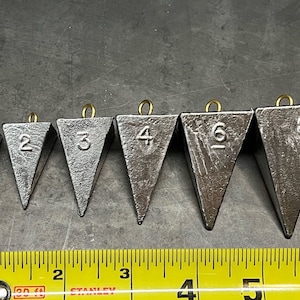 May include: A set of six triangular fishing sinkers, each with a brass loop and a number from 1 to 8 printed on the side. The sinkers are arranged in order of size, with the smallest sinker on the left and the largest sinker on the right. The sinkers are all made of lead and have a gray color.