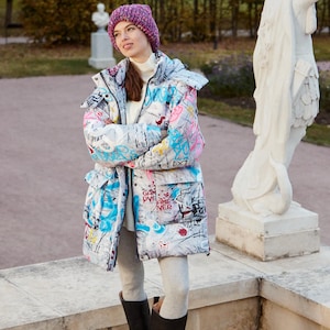 Peut inclure: Une femme portant une veste matelassée blanche et grise de style graffiti avec un bonnet rose, un legging gris et des bottes noires montant au genou. Elle se tient devant une statue de pierre blanche dans un parc.