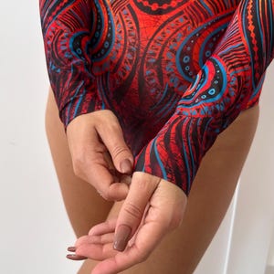 May include: A long-sleeved bodysuit with a vibrant, intricate pattern in red, black, and blue. The design features swirling shapes and geometric elements. The fabric appears to be a lightweight, stretchy material.