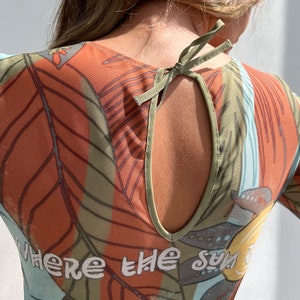 Puede incluir: Una blusa de malla transparente de manga larga, naranja, verde y azul, con estampado de hojas tropicales y el texto "Where the sun is" impreso en la parte delantera. La blusa tiene un escote en la espalda con un lazo verde para atar.
