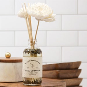 Puede incluir: Un frasco de vidrio transparente para difusor de varillas con l&iacute;quido y capullos de lavanda secos. La etiqueta dice MILK RECLAMATION BARN ESTD 2016 REED DIFFUSER LAVENDER LINEN NET WT 3.7 OZ. Dos flores de madera blanca sobresalen del cuello.