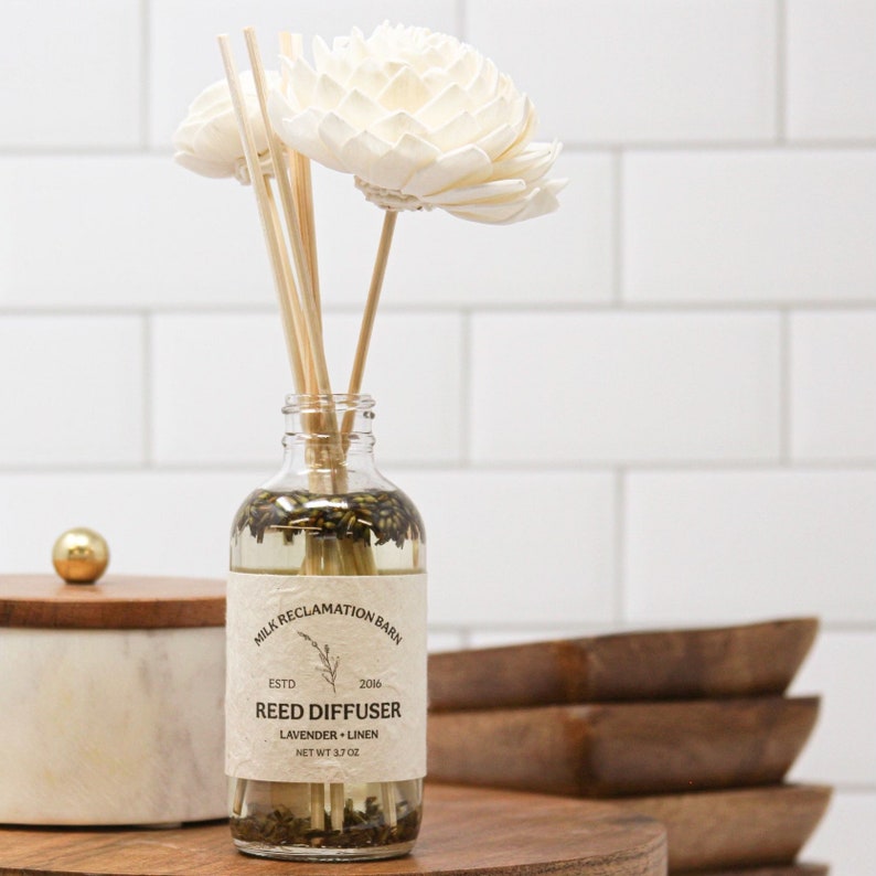 Puede incluir: Un frasco de vidrio transparente para difusor de varillas con l&iacute;quido y capullos de lavanda secos. La etiqueta dice MILK RECLAMATION BARN ESTD 2016 REED DIFFUSER LAVENDER LINEN NET WT 3.7 OZ. Dos flores de madera blanca sobresalen del cuello.