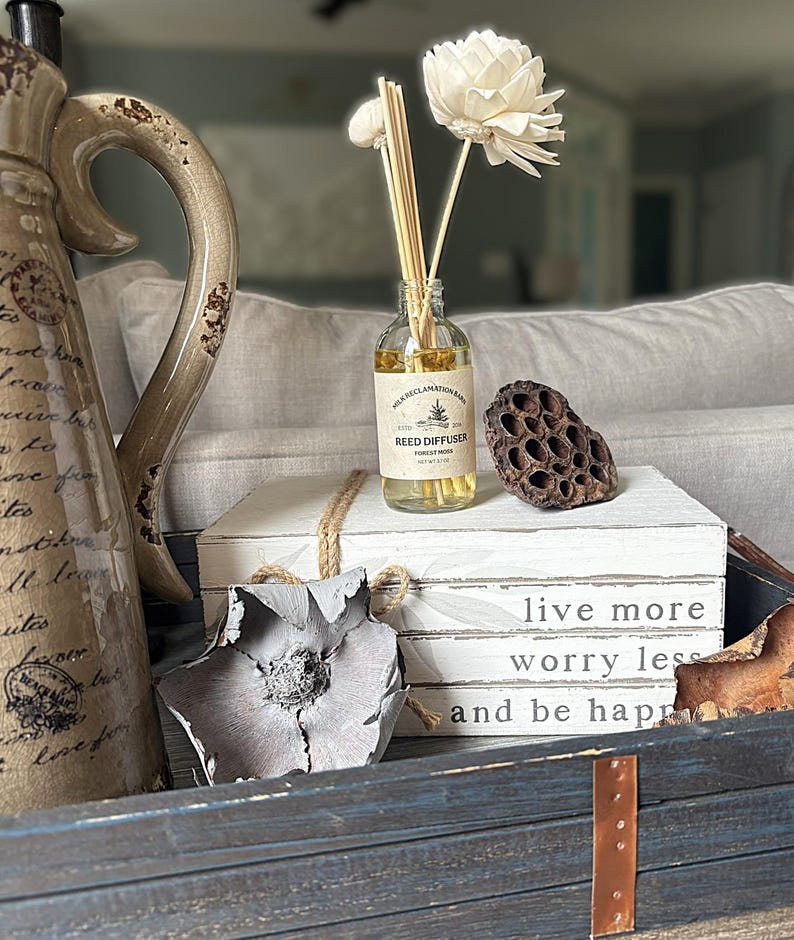 Puede incluir: Un frasco difusor de varillas de vidrio transparente con una etiqueta Milk Reclamation Barn Reed Diffuser Forest Moss, que contiene aceite amarillo claro y varillas de madera con una flor de madera sola blanca, sobre tres libros blancos.
