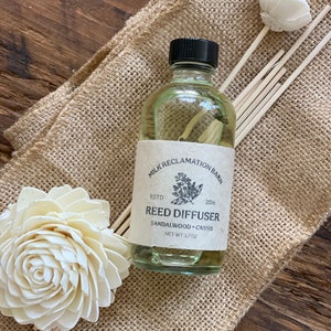 Puede incluir: Un frasco difusor de varillas de vidrio transparente con tapa negra y etiqueta beige que dice Milk Reclamation Barn, Estd 2016, Reed Diffuser, Sandalwood + Cassis, Net Wt 3.7 oz. Se acompa&ntilde;a de varias varillas de madera y dos flores blancas.