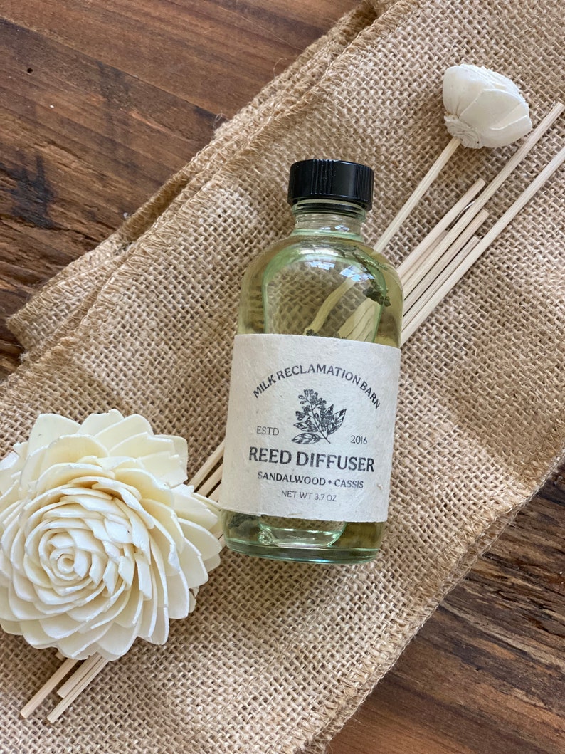 Puede incluir: Un frasco difusor de varillas de vidrio transparente con tapa negra y etiqueta beige que dice Milk Reclamation Barn, Estd 2016, Reed Diffuser, Sandalwood + Cassis, Net Wt 3.7 oz. Se acompa&ntilde;a de varias varillas de madera y dos flores blancas.