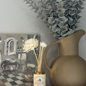 Puede incluir: Un frasco difusor de varillas de vidrio transparente con una etiqueta beige que dice Milk Reclamation Barn Reed Diffuser Forest Moss, que contiene l&iacute;quido &aacute;mbar claro y varillas de madera rematadas con dos flores de madera sola de color crema.