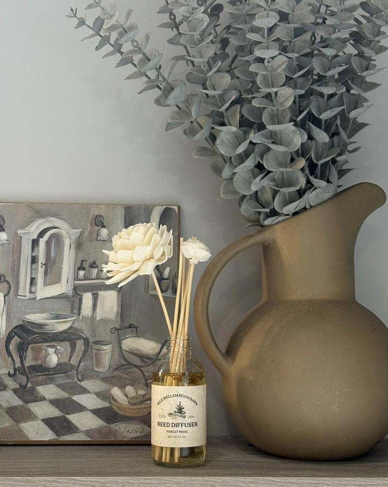 Puede incluir: Un frasco difusor de varillas de vidrio transparente con una etiqueta beige que dice Milk Reclamation Barn Reed Diffuser Forest Moss, que contiene l&iacute;quido &aacute;mbar claro y varillas de madera rematadas con dos flores de madera sola de color crema.