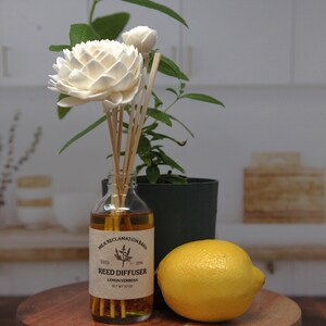 Puede incluir: Un frasco difusor de varillas de vidrio transparente lleno de l&iacute;quido &aacute;mbar, con una etiqueta que dice Milk Reclamation Barn, Reed Diffuser, Lemon Verbena, Net Wt 3.7 oz. Incluye varillas de madera rematadas con flores de madera sola blancas.