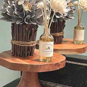Può includere: Un diffusore a bastoncini in una bottiglia di vetro trasparente con un'etichetta che riporta Milk Reclamation Barn Reed Diffuser Forest Moss Net Wt 3.7 oz, accompagnato da una composizione decorativa di fiori grigi e bianchi in un vaso di rametti.