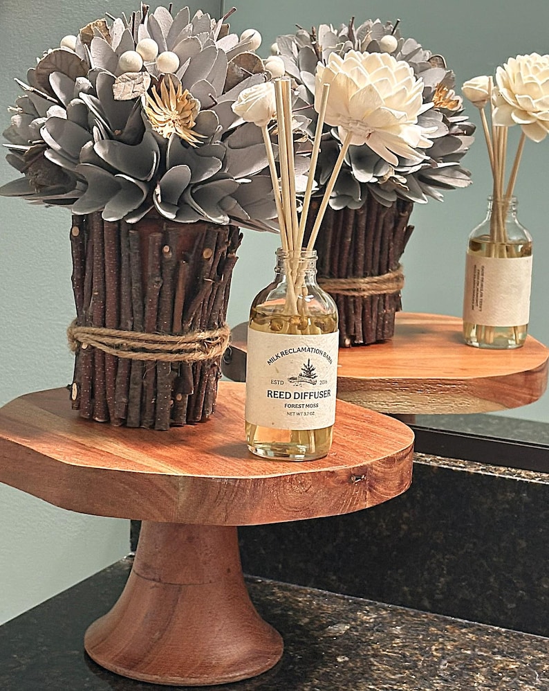 Puede incluir: Un difusor de varillas en una botella de vidrio transparente con una etiqueta que dice Milk Reclamation Barn Reed Diffuser Forest Moss Net Wt 3.7 oz, acompa&ntilde;ado de un arreglo decorativo de flores grises y blancas en un recipiente de ramas marrones.