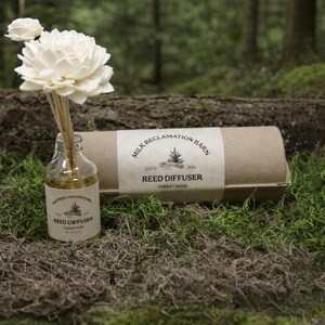 Puede incluir: Un frasco difusor de varillas de vidrio transparente con una etiqueta beige que dice Milk Reclamation Barn Estd 2016 Reed Diffuser Forest Moss, con flores de madera blancas. A su lado hay un tubo de cart&oacute;n cil&iacute;ndrico con la misma etiqueta.