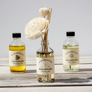 Puede incluir: Tres botellas de vidrio transparente con tapas negras y etiquetas beige que dicen Milk Reclamation Barn Reed Diffuser. La botella central contiene varillas de madera rematadas con dos flores de madera de sola de color crema.