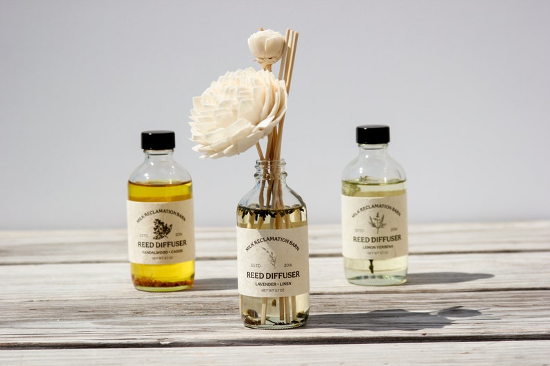 Puede incluir: Tres botellas de vidrio transparente con tapas negras y etiquetas beige que dicen Milk Reclamation Barn Reed Diffuser. La botella central contiene varillas de madera rematadas con dos flores de madera de sola de color crema.