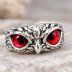 Peut inclure: Une bague en métal argenté représentant un visage de hibou détaillé avec des motifs de plumes texturés et deux yeux ronds en verre rouge vif proéminents insérés au centre de l'anneau, créant une apparence audacieuse et saisissante.