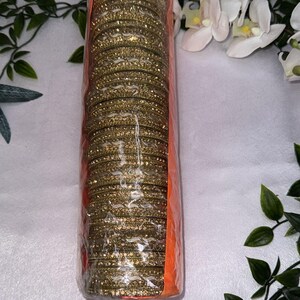 May include: A bundle of gold-coloured bangles wrapped in clear plastic and an orange backing. The bangles are covered in glitter and are stacked together. The background includes white flowers and green leaves.